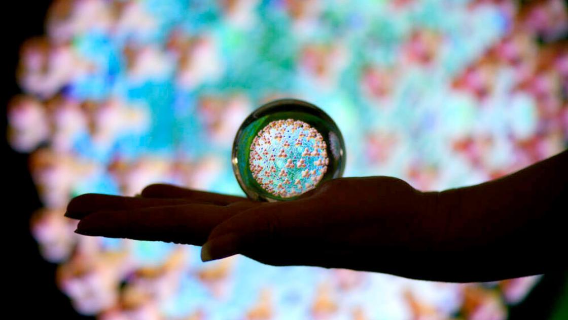 imago images / MiS Das Coronavirus gesehen durch eine Glaskugel auf einer Frauenhand.