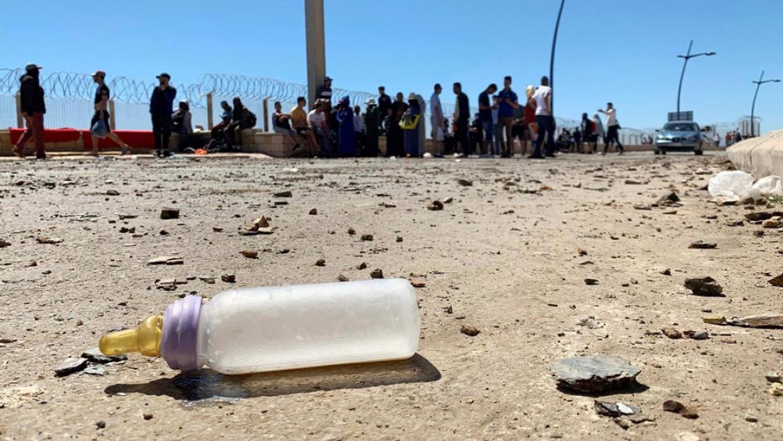 Eine Babyflasche auf dem Boden neben einer Gruppe von Menschen, die von der marokkanischen Bereitschaftspolizei vom Grenzübertritt zwischen Fnidq (Castillejos) und dem Grenzübergang Tarajal in Richtung der spanischen Stadt Ceuta in Nordafrika abgehalten wurden, 19. Mai 2021 (IMAGO / Agencia EFE)