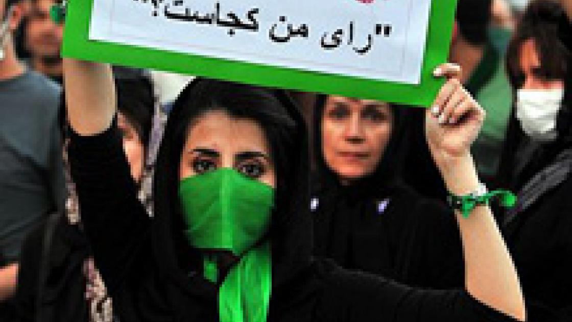 Das Bild zeigt eine schwarz gekleidete Frau mit einem grünen Tuch vor dem Mund. Sie hält ein Plakat in der Hand auf dem neben Schrift zwei rote Handabdrücke zu sehen sind