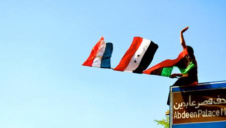 Das Bild zeigt eine Person, die die Flaggen von Lybien, Syrien und eine rot-weiß-blaue Flagge in Cairo weht